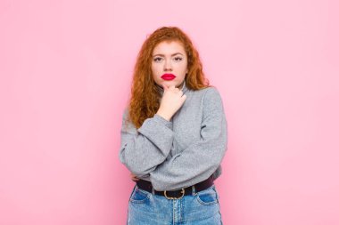 Genç kızıl saçlı kadın ciddi, şaşkın, kararsız ve düşünceli görünüyor. Seçenekler arasında veya pembe duvara karşı seçimler yapıyor.