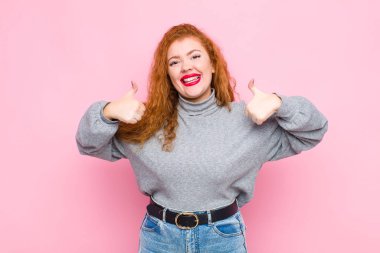 young red head woman smiling broadly looking happy, positive, confident and successful, with both thumbs up against pink wall clipart