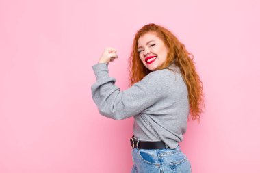 Genç kızıl saçlı kadın kendini mutlu, tatmin olmuş ve güçlü hissediyor, kaslı ve formda, spor salonundan sonra pembe duvara karşı güçlü görünüyor.