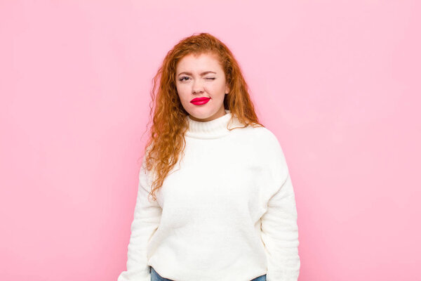 young red head woman looking happy and friendly, smiling and winking an eye at you with a positive attitude against pink wall