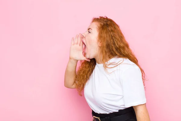 一个红头发的年轻女人生气地大声喊着要抄袭侧边的空间 手放在嘴边靠着粉红的墙壁 — 图库照片