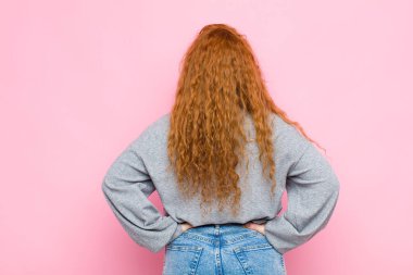 Genç kızıl saçlı kadın şaşkın ya da dolu hissediyor ya da şüphe ve sorular, merak ediyor, elleri kalçasında, pembe duvara bakan dikiz aynası