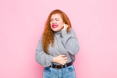 Genç kızıl saçlı kadın stresli, hayal kırıklığına uğramış ve yorgun hissediyor. Pembe duvara karşı endişeli ve sorunlu bir bakışla boynunu ovalıyor.