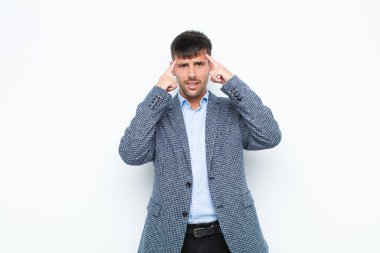 Genç ve yakışıklı bir adam ciddi ve konsantre görünümlü, beyin fırtınası yapıyor ve beyaz duvara karşı meydan okuyucu bir sorun düşünüyor.