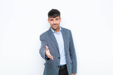 Genç yakışıklı adam gülümsüyor, mutlu, kendinden emin ve arkadaş canlısı görünüyor, anlaşmayı bitirmek için el sıkışmayı teklif ediyor, beyaz duvara karşı işbirliği yapıyor.