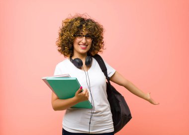 Genç ve güzel bayan öğrenci kendini mutlu ve neşeli hissediyor, gülümsüyor ve hoş geldiniz diyor, pembe duvara karşı dostça bir jestle sizi içeri davet ediyor.