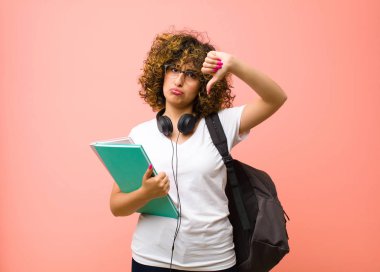 Genç, güzel bayan öğrenci üzgün, hayal kırıklığına uğramış ya da kızgın görünüyor. Anlaşmazlık içinde başparmaklarını gösteriyor. Pembe duvara karşı hayal kırıklığına uğramış hissediyor.