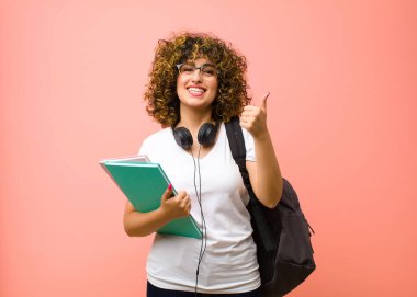 Genç ve güzel bayan öğrenci neşeyle gülümsüyor ve mutlu görünüyor, kaygısız ve pozitif hissediyor iki başparmağı pembe duvara dayalı