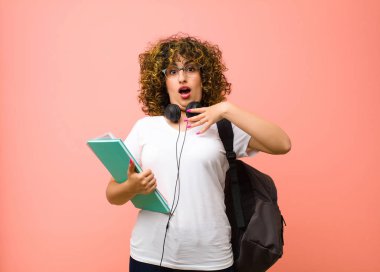 Genç ve güzel bayan öğrenci şok olmuş, şaşırmış ve şaşırmış hissediyor, elleri göğsünde ve ağzı açık, kim diyor, ben mi? Pembe duvara yaslan