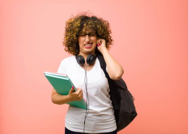 Genç, güzel bir bayan öğrenci sinirli, stresli ve sinirli görünüyor, her iki kulağı da sağır edici bir ses, ses veya pembe duvara karşı yüksek sesle müzik dinliyor.
