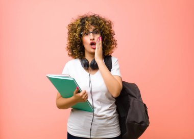 Genç ve güzel bir bayan öğrenci şok olmuş ve korkmuş hissediyor. Ağzı açık ve elleri pembe duvarda.