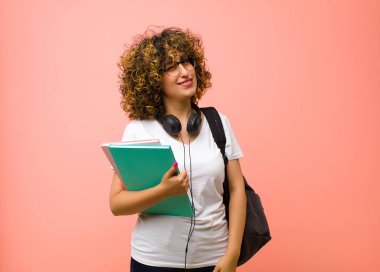 Genç ve güzel bayan öğrenci mutlu ve arkadaş canlısı görünüyor, pembe duvara karşı pozitif bir tavırla sana gülümsüyor ve göz kırpıyor.