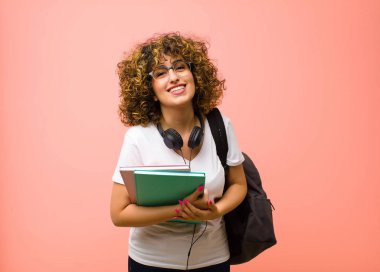 Genç ve güzel bir bayan öğrenci, neşeli bir şekilde gülümsüyor ve kollarını kavuşturup kollarını kavuşturuyor, pembe duvara karşı mutlu ve pozitif hissediyor.