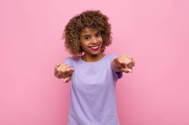 Genç Afrikalı Amerikalı kadın mutlu ve kendinden emin, kamerayı iki eliyle gösteriyor ve gülüyor, pembe duvara karşı seni seçiyor.