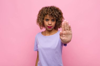 Genç Afrikalı Amerikalı kadın ciddi, sert, hoşnutsuz ve kızgın görünüyor. Pembe duvara karşı el hareketini durduruyor.