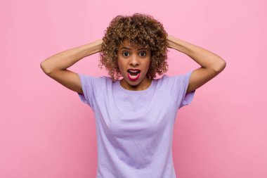 Genç Afrikalı Amerikalı kadın kendini stresli, endişeli ya da korkmuş hissediyor. Elleri başının üstünde, pembe duvara karşı panikliyor.