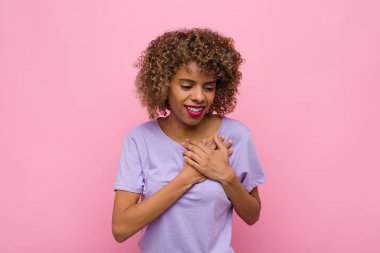 Genç Afrikalı Amerikalı kadın üzgün, yaralı ve kalbi kırık görünüyor, iki elini de kalbe yakın tutuyor, ağlıyor ve pembe duvara karşı depresyona giriyor.