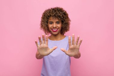 Genç Afrikalı Amerikalı kadın gülümsüyor ve arkadaş canlısı görünüyor, onuncu ya da onuncu numarayı gösteriyor, elleri önde, pembe duvara doğru sayıyor.