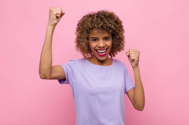 Genç Afrikalı Amerikalı kadın muzaffer bir şekilde bağırıyor. Heyecanlı, mutlu ve şaşırmış görünüyor. Pembe duvara karşı kutlama yapıyor.