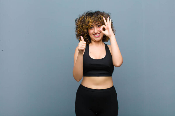 young sports woman feeling happy, amazed, satisfied and surprised, showing okay and thumbs up gestures, smiling against gray wall