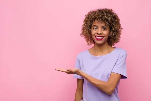 若いアフリカ系アメリカ人の女性が陽気に笑って幸せを感じながらピンクの壁に手のひらでコピースペースでコンセプトを示し — ストック写真