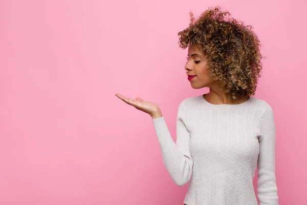 young african american woman feeling happy and smiling casually, looking to an object or concept held on the hand on the side against pink wall