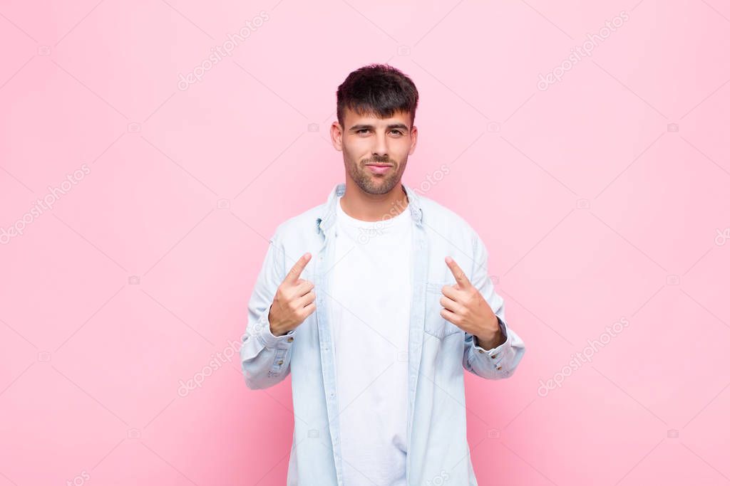 young handsome man with a bad attitude looking proud and aggressive, pointing upwards or making fun sign with hands against pink wall
