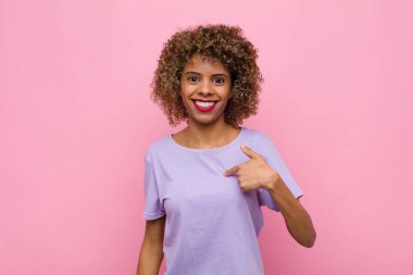 Genç Afrikalı Amerikalı kadın mutlu, gururlu ve şaşırmış görünüyor. Neşeli bir şekilde kendini gösteriyor. Kendine güveniyor ve pembe duvara karşı kibirli.