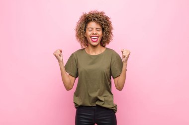 Genç Afro-Amerikalı kadın son derece mutlu ve şaşırmış görünüyor, başarıyı kutluyor, bağırıyor ve pembe duvara zıplıyor.
