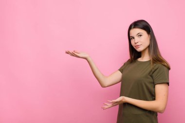 Genç ve güzel bir kadın gururla ve güvenle gülümsüyor, mutlu ve tatmin olmuş hissediyor ve pembe duvara karşı fotokopi alanında bir konsept gösteriyor.