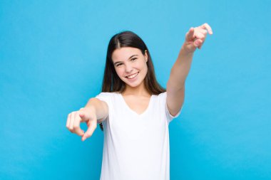 Genç ve güzel bir kadın mutlu ve kendinden emin, kamerayı iki eliyle gösteriyor ve gülüyor, seni mavi duvara karşı seçiyor.