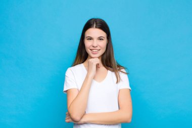 Genç ve güzel bir kadın mutlu görünüyor ve gülümsüyor. Elleri çenesinde, merak ediyor ya da soru soruyor. Mavi duvarla seçenekleri karşılaştırıyor.