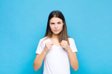 Genç ve güzel bir kadın kendinden emin, sinirli, güçlü ve agresif görünüyor. Yumrukları mavi duvara karşı boks pozisyonunda dövüşmeye hazır.