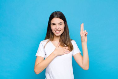 Genç, güzel bir kadın mutlu, kendinden emin ve güvenilir görünüyor. Gülümsüyor ve zafer işareti gösteriyor. Mavi duvara karşı olumlu bir tutum sergiliyor.