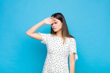 Genç ve güzel bir kadın stresli görünüyor, yorgun ve hayal kırıklığına uğramış, alnından ter damlıyor, mavi duvara karşı umutsuz ve bitkin hissediyor.