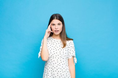 Genç, güzel bir kadın şaşırmış, ağzı açık, şok olmuş, mavi duvara karşı yeni bir düşünce, fikir ya da konsept fark ediyor.