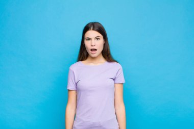 Genç ve güzel bir kadın korkmuş ve şok olmuş hissediyor, ağzı açık mavi duvara karşı şaşkınlık içinde.