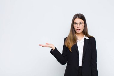 Genç iş kadını şaşırmış ve şoka uğramış görünüyor, çenesi düşmüş, beyaz duvara karşı eli açık bir obje tutuyor.