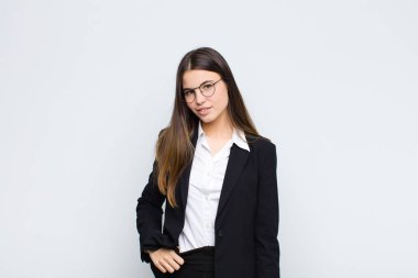 Genç iş kadını beyaz duvara karşı neşeli, mutlu, kendinden emin ve rahat bir ifadeyle gülümsüyor.
