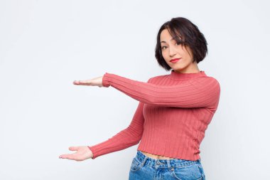 Genç ve güzel bir kadın, iki eli de kenarda olan bir objeyi tutuyor. Fotokopi alanı, beyaz duvara karşı bir objeyi gösteriyor, sunuyor ya da reklamını yapıyor.