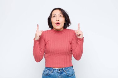 Genç ve güzel bir kadın şaşkınlık ve ağzı açık bir şekilde beyaz duvara karşı şaşkın ve şaşkın bir bakışla yukarıya bakıyor.