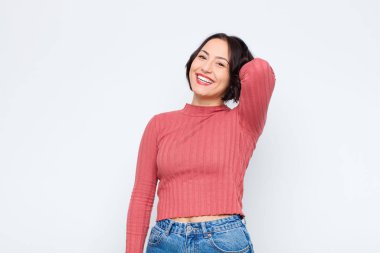 Genç, güzel bir kadın neşeli ve tesadüfen gülümsüyor, beyaz duvara karşı pozitif, mutlu ve kendinden emin bir bakışla göğüs göğüse tutuşuyor.