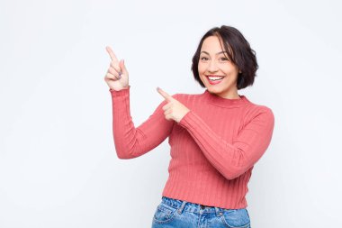 Genç, güzel bir kadın sevinçli ve şaşırmış hissediyor. Şok olmuş bir ifadeyle gülümsüyor ve beyaz duvara doğru işaret ediyor.