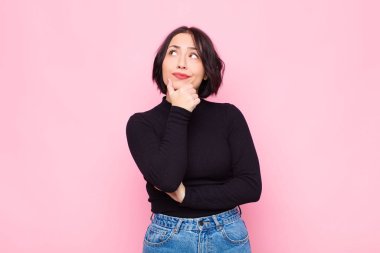 Genç, güzel bir kadın düşünüyor, kuşkulu ve şaşkın hissediyor, farklı seçeneklerle, pembe duvara karşı hangi kararı vereceğini merak ediyor.