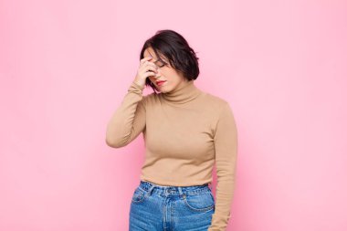 Genç ve güzel bir kadın stresli, mutsuz ve hayal kırıklığına uğramış, alnına dokunuyor ve pembe duvara karşı şiddetli baş ağrısı çekiyor.