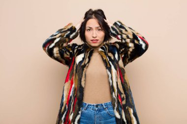 Genç, güzel bir kadın kendini stresli, endişeli, endişeli ya da korkmuş hissediyor. Elleri başının üstünde, bej rengi duvara karşı panikliyor.