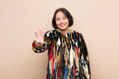 Genç, güzel bir kadın gülümsüyor ve arkadaş canlısı görünüyor. Üç ya da üç numarayı gösteriyor. Elleri önde, bej rengi duvara doğru sayıyor.
