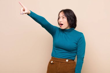 Genç, güzel bir kadın şok olmuş ve şaşırmış hissediyor. Bej duvara karşı hayret dolu, açık ağızlı bir bakışla yukarılara bakıyor.