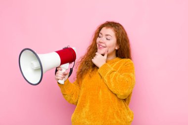 Genç kızıl saçlı kadın mutlu, kendinden emin bir ifadeyle gülümsüyor. Çenesinde elleri, merak ediyor ve megafonla yan tarafa bakıyor.