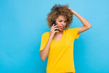 Genç Afro-Amerikan duyguları şaşkın ve kafası karışmış, kafasını kaşıyor ve cep telefonuyla yan tarafa bakıyor.